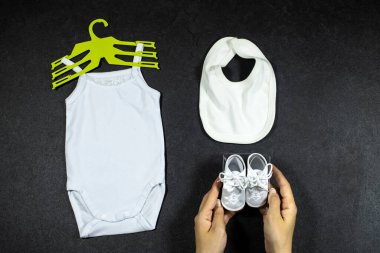 White baby girl or boy bodysuit mockup flat lay with bib and hand hold shoes on grunge gray concrete background. Design onesie template, print presentation mock up