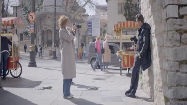 Woman photographs young man in old European city. Action. Side view of woman photographing man with camera. Shooting man on professional camera in old town — Stock Video