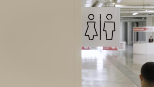 Los baños de hombres y mujeres firman en el centro comercial. HDR. Vista trasera de un hombre de camisa blanca caminando por la pared beige con símbolos masculinos y femeninos de un baño público. — Vídeos de Stock