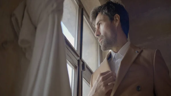 El hombre de traje está de pie junto a la ventana. Acción. El hombre elegante en traje de luz está de pie junto a la ventana. Hombre atractivo en traje elegante se encuentra en la ventana en la antigua mansión o castillo — Foto de Stock