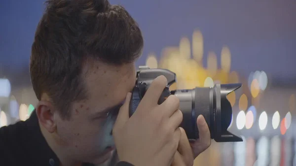 Jeune homme prend des photos avec un appareil photo sur fond de ville nocturne. L'action. Beau jeune homme avec caméra professionnelle sur fond de ville floue en soirée — Photo