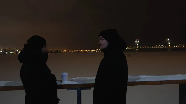 两个年轻人坐在一条冰冻的河边，聊得很愉快。概念。两名男子站在白雪覆盖的池塘边，背后是城市的灯光和漆黑的天空. — 图库照片
