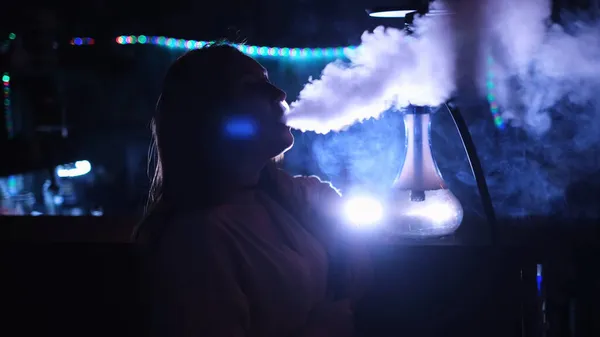 Close up vista lateral de uma menina fumando narguilé em fundo de néon azul. Mídia. Feminino enquanto fuma um narguilé em um clube, exalando uma nuvem de névoa e refrigeração. — Fotografia de Stock
