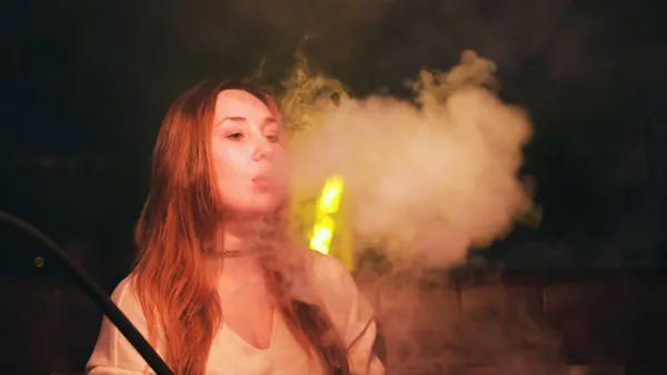 Retrato de uma mulher fumando cachimbo de água em uma sala de estar. Mídia. Mulher com cabelo vermelho longo e gargantilha preta no pescoço relaxando em um bar shisha. — Fotografia de Stock