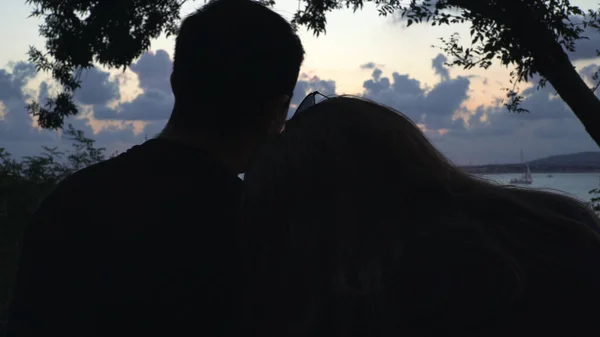 Couple of lovers sitting together on beach cost and looking far at ocean horizon and sunset sky. Media. Boy and girl silhouettes embracing at sea with her head on his shoulder. — Stock Photo, Image