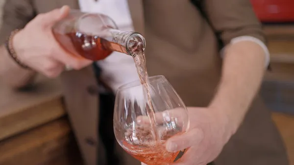 Primo piano di un uomo in abito beige che versa vino rosato in un bicchiere trasparente. Azione. Sommelier maschile versando vino di frutta in un bar. — Foto Stock