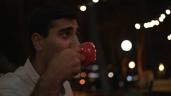 Joven hombre guapo sentado afuera en la cafetería de la ciudad y bebiendo bebida caliente de una gran taza roja. Medios. Hombre bebiendo café en la noche en el cielo negro y fondo de guirnalda. — Foto de Stock