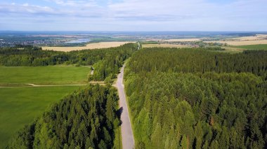 Helikopterden görüntü. Yaratıcı. Yazın yeşil bir orman, bir yol, şehrin küçük bir kısmı, arkasında mavi bir gökyüzü, bir nehir ve küçük bir tarla görünür..