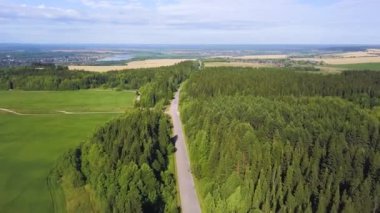 Helikopterden görüntü. Şarjör. Yazın yeşil bir orman, bir yol, şehrin küçük bir kısmı, arkasında mavi bir gökyüzü, bir nehir ve küçük bir tarla görünür..