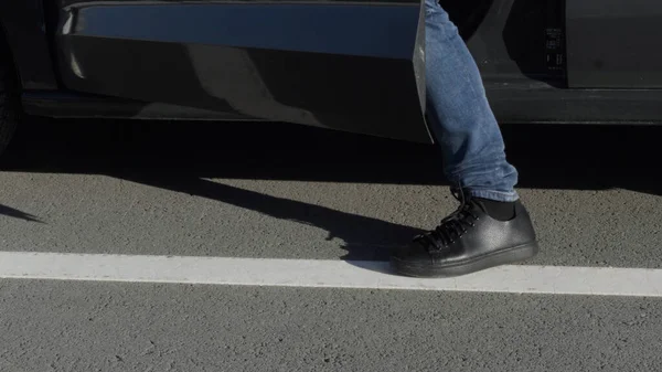 Чоловік сідає в машину на стороні водіїв. HDR. Крупним планом чоловічі ноги в чорному взутті і джинсах на фоні асфальтової дороги і легкового автомобіля . — стокове фото