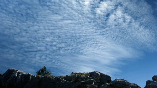 Un cielo claro ligeramente nublado con un patrón. Creativo. Sobre el fondo del cielo hay una hermosa montaña gris con un pequeño número de árboles y piedras grises debajo.. —  Fotos de Stock