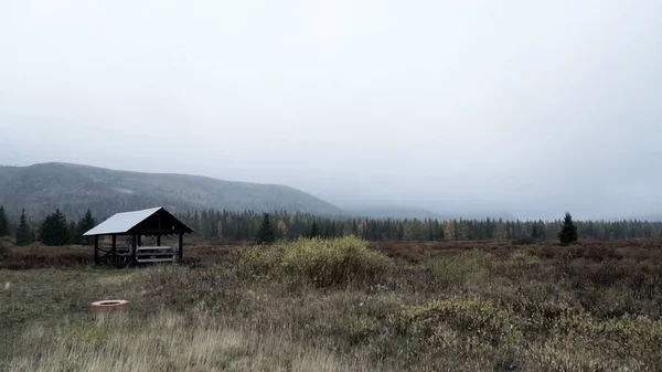 Een verlaten veld. Een knip. Gloomy ochtendlandschap met een houten huis en sombere bergen op de achtergrond — Stockfoto