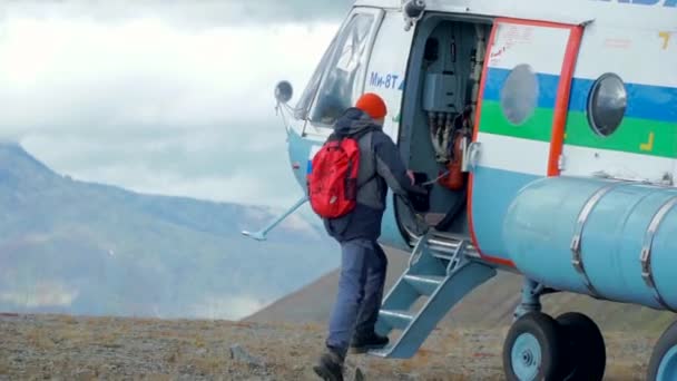 Subiendo a un helicóptero. Clip. Los hombres entran en un helicóptero que se encuentra en el bosque sobre el fondo de un cielo nublado y enormes montañas. — Vídeo de stock