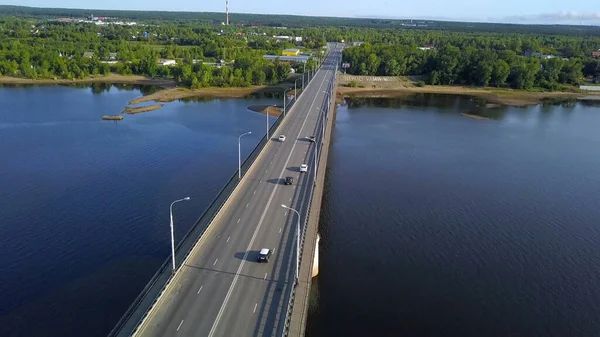 Vista desde la altura. Clip. Un gran puente largo sobre el río en el que los coches conducen y hay luces detrás del puente se puede ver una ciudad verde con pequeños edificios residenciales y un gran bosque —  Fotos de Stock