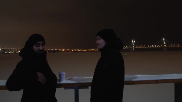 Due ragazzi vicino a un fiume ghiacciato hanno una bella conversazione. Concetto. Due uomini in piedi all'aperto vicino allo stagno innevato con le luci della città e il cielo nero sullo sfondo. — Video Stock