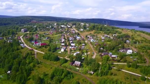View from the height . Clip . A small summer village with a river, small residential houses and roads, a green forest with large trees and hills is visible from behind. — Stock Video