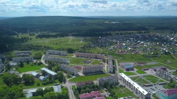 Vista desde un helicóptero. Clip. Una pequeña ciudad verde de verano con parques y casas para la gente, un gran bosque y colinas con campos verdes son visibles desde atrás — Vídeos de Stock