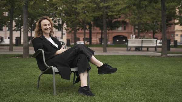 Porträt einer schönen Frau mittleren Alters mit lockigem Haar, die im Stadtpark auf einem Stuhl sitzt. Handeln. Glücklich lachende Frau in schwarzer Kleidung sitzt mit schwarzer Kamera in der Hand. — Stockfoto