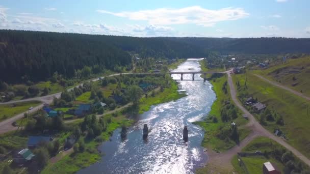 Uma paisagem aérea drone de uma pequena aldeia com rio e verdes montanhas arborizadas. Clipe. Uma ponte que cruza duas margens do rio no fundo do céu nublado. — Vídeo de Stock