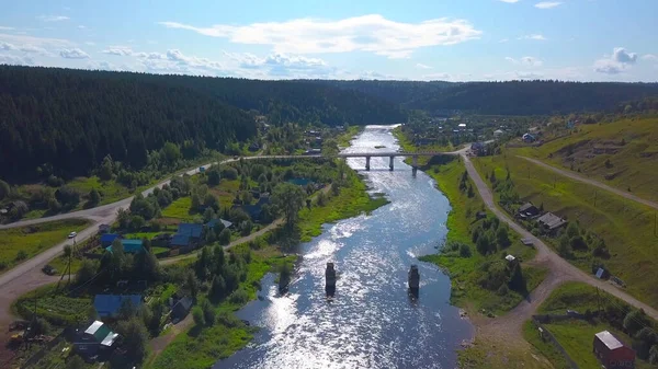 Eine Drohnenlandschaft aus der Luft, die ein kleines Dorf mit Fluss und grünen bewaldeten Bergen zeigt. Clip. Eine Brücke über zwei Flussufer bei bewölktem Himmel. — Stockfoto