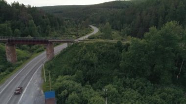 Virajlı yolun yukarısında terk edilmiş bir demiryolu köprüsü ve bir sürücü kamyonu. Sahne. Yeşil yaz ormanı ve asfalt köprünün altında destek sütunlarıyla yol bükülmüş..
