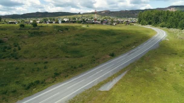 村に向かって緑のフィールドを介して実行されている空中田舎道。現場だ。青い空を背景に道路と小さな集落を曲げる. — ストック動画