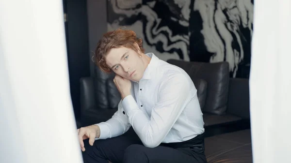 Un hombre con una camisa blanca. Acción. Un hombre con el pelo rojo está sentado en el sofá y ajustar el cuello de su ropa sobre un fondo de fondo de pantalla negro claro y una cortina blanca brillante en —  Fotos de Stock