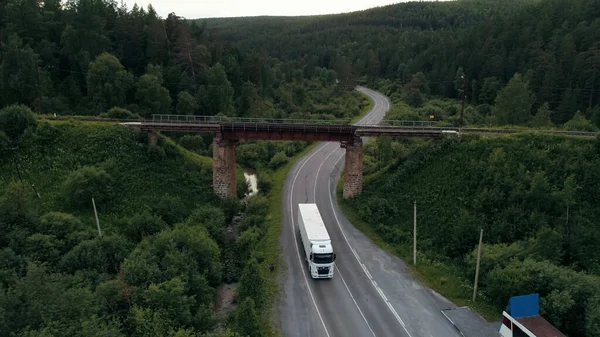 En övergiven rostig järnvägsbro ovanför den svängda vägen med en lastbil. På plats. Grön sommar skog och asfalt böjda väg under bron med stödjande kolumner. — Stockfoto