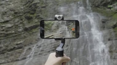 Dağları ve şelaleleri vuruyor. Başla. Telefon bir standın üzerinde duruyor ve dağları ve damlayan küçük bir şelaleyi olan güzel manzaraları çekiyor. .