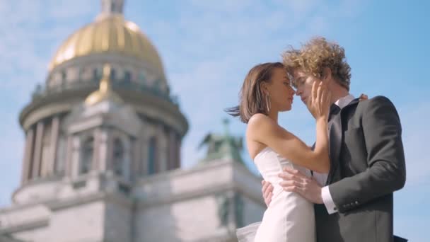 Sesión de fotos de boda. Acción. Una pareja posa junto a la Catedral de San Isaac, un joven con el pelo largo y rizado y su novia con hombros desnudos y pendientes largos — Vídeos de Stock