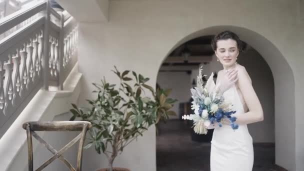 Жінка позує з квітами. Дія. Продумана молода наречена в розкішній довгій весільній сукні тримає букет і стоїть біля будинку зі сходами . — стокове відео