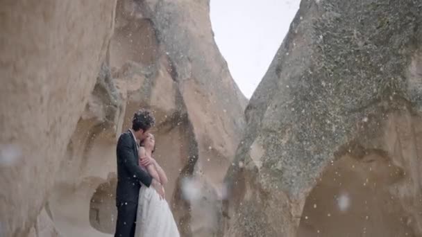 ロマンチックな雪の下でカッパドキアの山の中で美しい若い結婚式のカップル。行動だ。雪の結晶の下で珍しい岩の形成に囲まれた峡谷に一緒に立って花嫁と新郎. — ストック動画