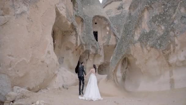 Hermosa joven pareja de boda en las montañas de Capadocia bajo una romántica nevada. Acción. Novia y novio juntos de pie en el barranco rodeado de formaciones rocosas inusuales bajo la caída de copos de nieve. — Vídeos de Stock