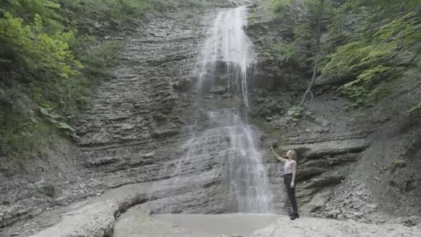 Žena fotí nebo natáčí video z hor a vodopádu na svém smartphonu pomocí stativu. Akce. Žena cestovatel s selfie hole a telefon stojící v rokli obklopen zelenou — Stock video