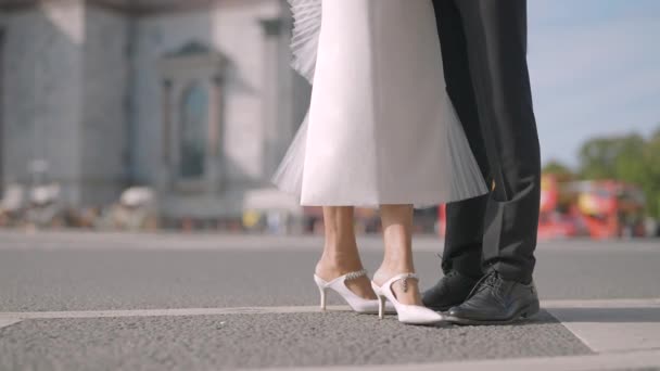 Legs of stylish bride and groom on the background of city summer street with driving cars. Action. Man in black suit and a woman in white dress. — Video Stock