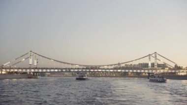 Işıklarla süslenmiş köprüden geçen güzel dalgalı nehir. Başla. Günbatımı arka planında geniş nehri geçen turistik yatlar.