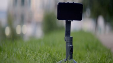 Modern akıllı telefon açık havada bir yaz gününde tripoda takılır. Başla. Bulanık şehir arka planında çayırda yeşil çimlerin üzerinde duran telefonu olan bir tripod..