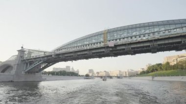 Nehirde cam köprüsü olan yüzen bir teknenin görüntüsü. Başla. Şehrin arka planında ve cam köprüde güzel bir nehir manzarası. Açık güneşli bir günde modern jöleli köprü