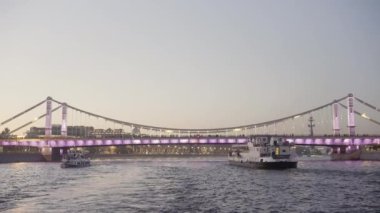 Işıklarla süslenmiş köprüden geçen güzel dalgalı nehir. Başla. Günbatımı arka planında geniş nehri geçen turistik yatlar.