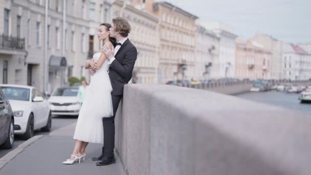 Vedere laterală de îngrijire frumos doar căsătorit cuplu îmbrățișarea de trotuar de granit. Acţiune. Mireasă elegantă și mire în aer liber în stradă. — Videoclip de stoc