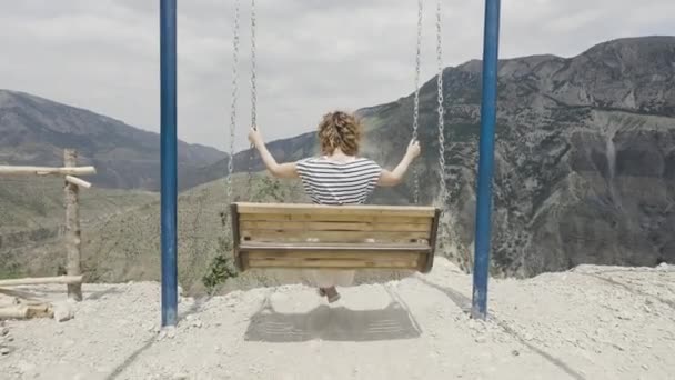 Rear view of a young woman swinging in mountains. Action. Concept of tourism, travel, and freedom swinging on the background of mountains and cloudy sky. — Video Stock