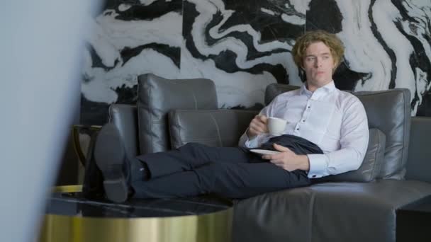 Um homem bonito com cabelo comprido está descansando. Ação.O belo noivo está sentado descansando e bebendo uma xícara de café no quarto no sofá. — Vídeo de Stock