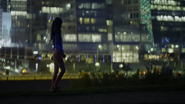 Beautiful girl goes on background of night city. Action. Girl with ponytails walks on background of panorama of modern night city. Lost girl on background of big night city — стоковое фото