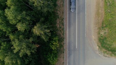 View from a helicopter . Scene . A narrow road on which a cargo truck with a blue front and a car with a trailer for transportation are traveling , there is a forest and a road with grass nearby .