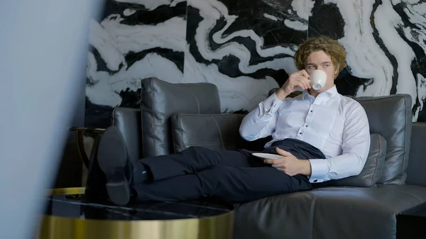 Un hombre guapo con el pelo largo está descansando. Acción.El guapo novio está sentado descansando y bebiendo una taza de café en la habitación en el sofá. — Foto de Stock