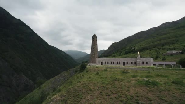 Kaukasischer Kampfturm, Inguschetien. Handeln. Luftaufnahme eines alten Turms und eines Gebäudes auf einem grünen Hügel, umgeben von Bergen. — Stockvideo