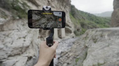 Dağları ve şelaleleri vuruyor. Başla. Telefon bir standın üzerinde duruyor ve dağları ve damlayan küçük bir şelaleyi olan güzel manzaraları çekiyor. .