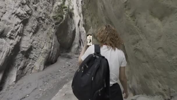 Вид спереду молода жінка, що йде в ущелині між каменями з телефоном у руках. Дія. Жіночий мандрівник, що знімає красиве утворення природного каменю . — стокове відео