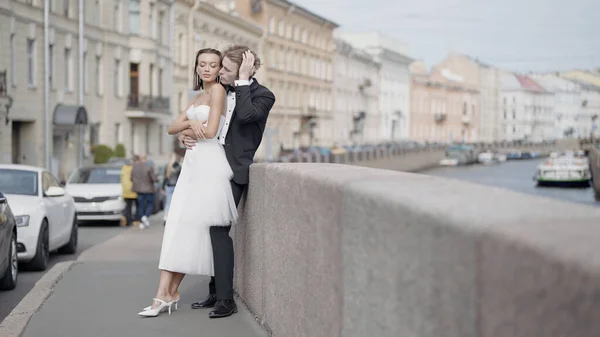 Όμορφοι νιόπαντροι. Πάμε. Ένα ζευγάρι των εραστών, η νύφη σε ένα σφιχτό λευκό φόρεμα και ο γαμπρός σε ένα κοστούμι με μακριά μαλλιά θέτουν στο δρόμο δίπλα στο ανάχωμα και όμορφα ιστορικά κτίρια στο — Φωτογραφία Αρχείου
