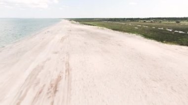 Kocaman bir sahil. Başla. Arkasında bitki örtüsü, yeşil tarlalar ve ağaçların olduğu deniz ve kumlu güzel bir sahil manzarası..